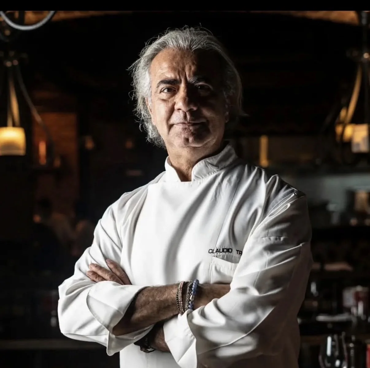 Ritratto intenso dello chef Claudio Trovato, a braccia conserte in un ristorante in penombra.