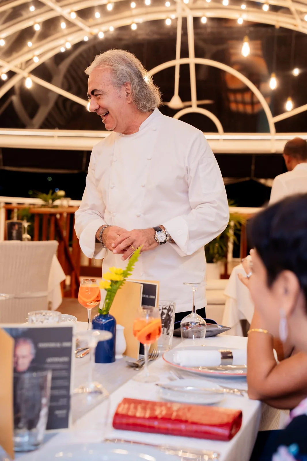 Lo chef ride con gli ospiti a una cena di gala, sotto le luci di un dehors elegante.